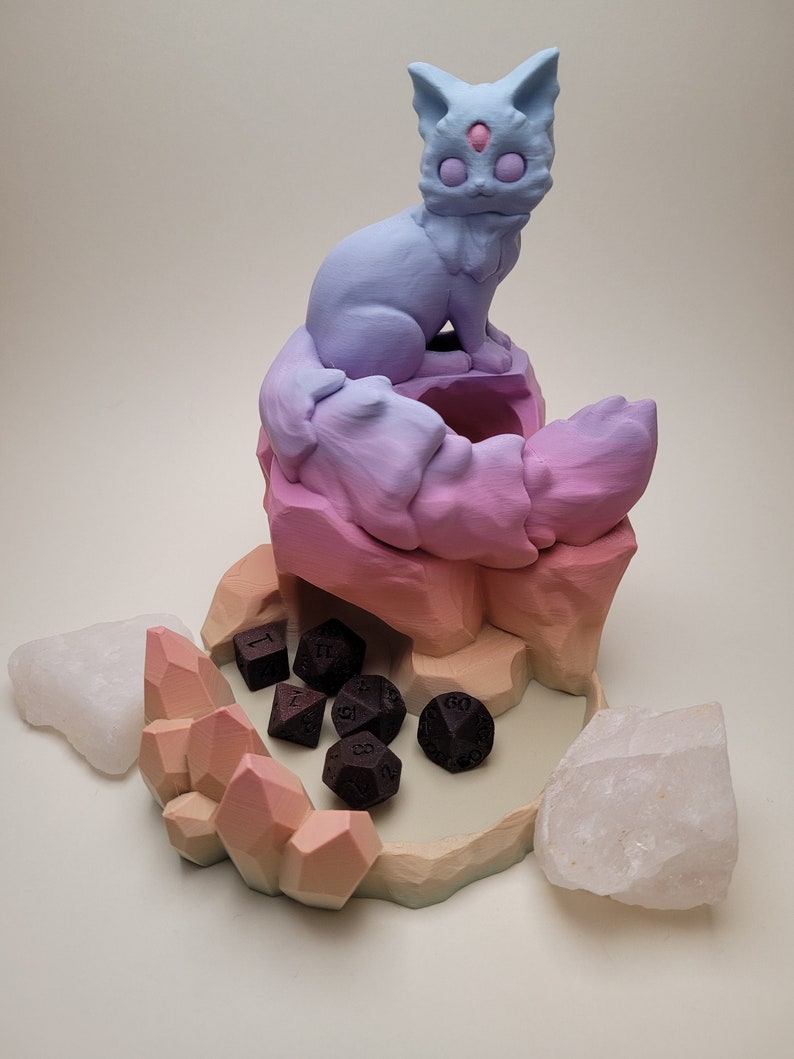 May include: A 3D printed dice tower with a pastel pink, purple, and blue gradient. A cat figurine sits on top of the tower. The tower has a crystal-shaped base and is surrounded by six black dice.