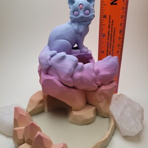 May include: A 3D printed figurine of a purple and blue cat sitting on a multi-colored rock formation with a crystal cave. The figurine is on a white surface.