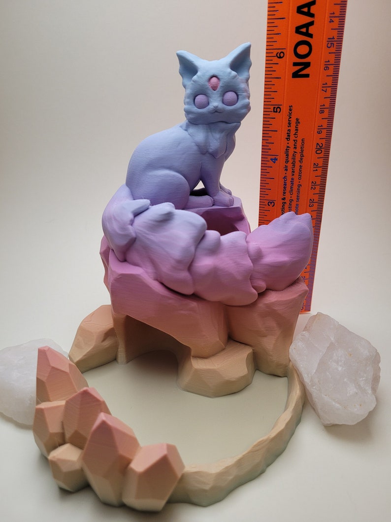 May include: A 3D printed figurine of a purple and blue cat sitting on a multi-colored rock formation with a crystal cave. The figurine is on a white surface.