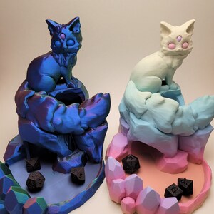 May include: Two 3D printed cat dice towers, one blue and one pink, with black dice inside. The towers are shaped like cats with crystal-like bases.