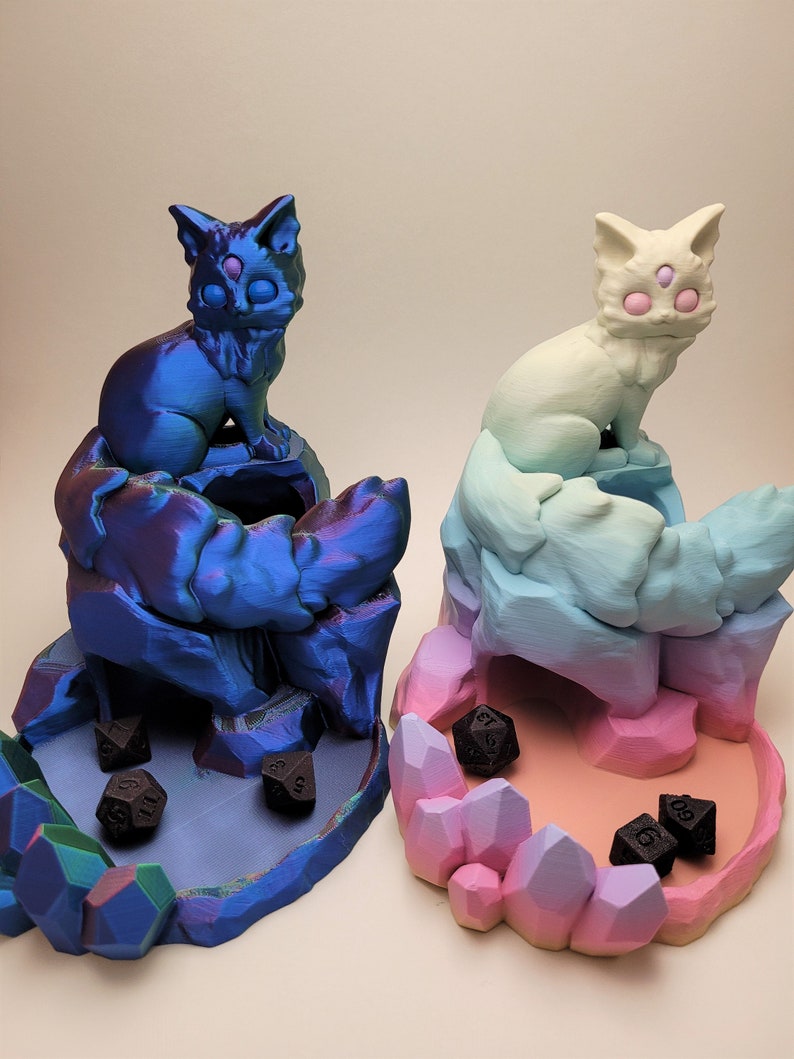 May include: Two 3D printed cat dice towers, one blue and one pink, with black dice inside. The towers are shaped like cats with crystal-like bases.