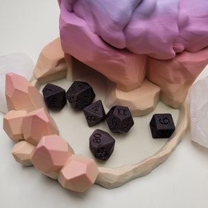 May include: A set of six black dice with white numbers, resting in a colorful, 3D printed dice tower. The tower is shaped like a mountain range with a pink, purple, and orange gradient.