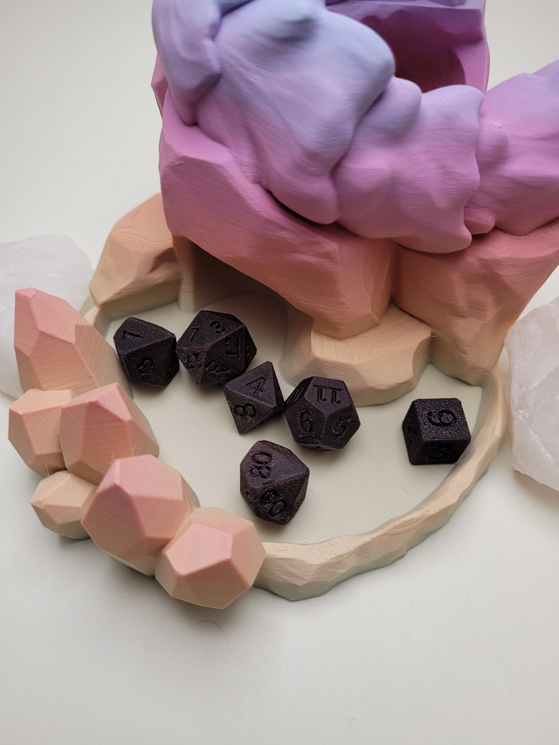 May include: A set of six black dice with white numbers, resting in a colorful, 3D printed dice tower. The tower is shaped like a mountain range with a pink, purple, and orange gradient.