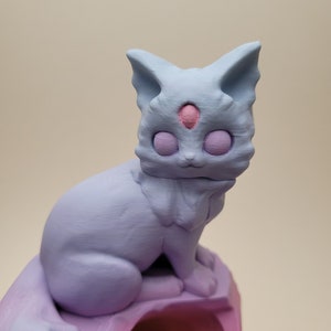 May include: A light blue, 3D printed figurine of a cat with a third eye in the center of its forehead. The cat is sitting on a pink, 3D printed base.