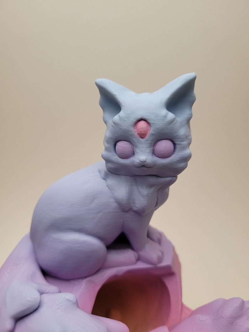 May include: A light blue, 3D printed figurine of a cat with a third eye in the center of its forehead. The cat is sitting on a pink, 3D printed base.