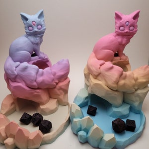 May include: Two 3D printed dice towers shaped like mountains with a cat on top. The towers are painted in a gradient of pink, orange, and yellow. The cat on the left tower is blue and the cat on the right tower is pink. Both cats have three eyes. There are black dice in the bottom of each tower.