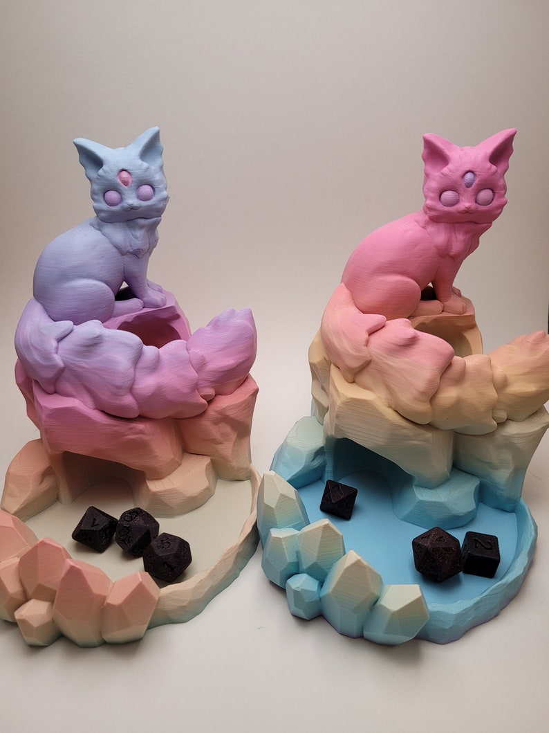 May include: Two 3D printed dice towers shaped like mountains with a cat on top. The towers are painted in a gradient of pink, orange, and yellow. The cat on the left tower is blue and the cat on the right tower is pink. Both cats have three eyes. There are black dice in the bottom of each tower.