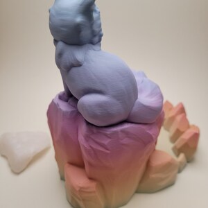 May include: A 3D printed figurine of a cat sitting on a rock formation. The cat is light blue and the rock is a gradient of pink, orange, and yellow.