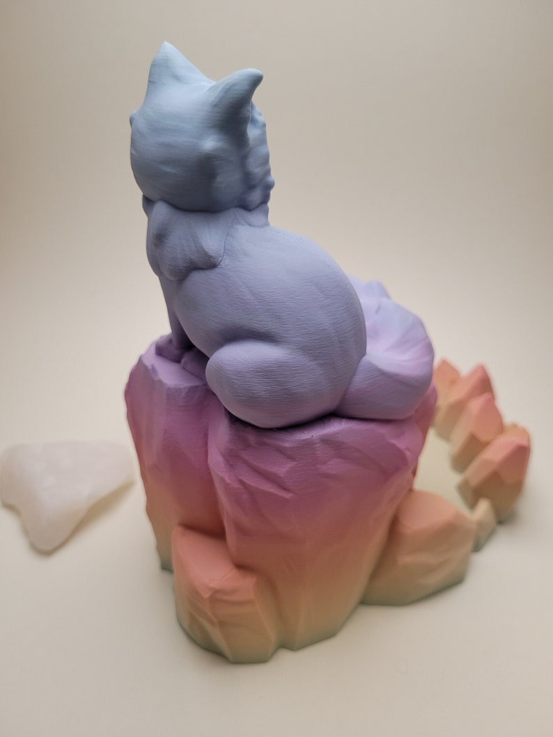 May include: A 3D printed figurine of a cat sitting on a rock formation. The cat is light blue and the rock is a gradient of pink, orange, and yellow.