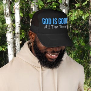 Puede incluir: Una gorra de estilo camionero negra con parte trasera de malla y panel frontal sólido. El frente muestra el texto "GOD IS GOOD" en letras grandes de color azul brillante, con la frase "All The Time" impresa debajo en letras blancas texturizadas.