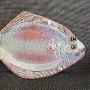 May include: A ceramic fish-shaped serving dish with a textured, glazed finish. The dish features a tail and fins, with a color palette of white, red, and blue. The dish measures approximately 12 inches long.