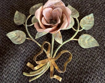 May include: A metal wall art piece shaped like a rose with a central pinkish-copper flower, green-toned leaves, and a gold-colored ribbon tied around the stems. The object features a weathered, textured finish across the petals and foliage.