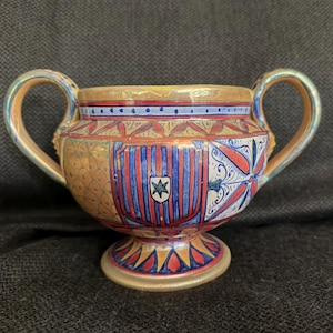 May include: A decorative ceramic bowl with two handles, featuring a detailed painted design. The bowl has a gold rim and base, with a central design of red and blue stripes and a star emblem. The handles are curved and painted in shades of gold and blue.