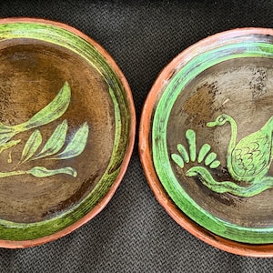 May include: Two handmade ceramic plates with brown centers and green rims. One plate features a green bird and foliage design, the other a green swan. The plates have a rustic, earthy aesthetic.