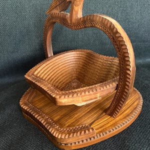 May include: A wooden apple-shaped fruit basket with a carved leaf detail. The basket is made of light brown wood and features a folding design. The base and handle are also carved with decorative patterns.
