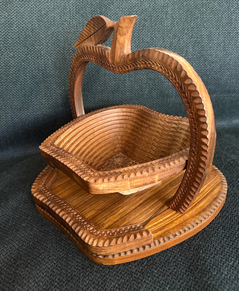 May include: A wooden apple-shaped fruit basket with a carved leaf detail. The basket is made of light brown wood and features a folding design. The base and handle are also carved with decorative patterns.