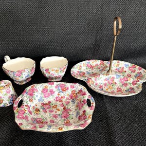 May include: A porcelain tea set featuring a vibrant floral pattern with pink roses and small blue and yellow flowers. The set includes two teacups, one small bowl, a square dish with two side handles, and a three-sectioned serving tray with a metal handle.