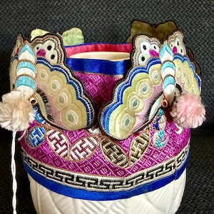 May include: A vibrant magenta fabric headpiece featuring intricate embroidery of butterflies with light blue and green wings. The base is adorned with a geometric Greek key border in black and white, accented by small pink pom-poms on each side.