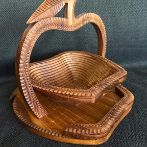 May include: A wooden apple-shaped basket with a carved leaf detail. The basket has a layered design and sits on a matching base. The wood has a warm, natural tone, and the basket is likely used for decorative or functional purposes.
