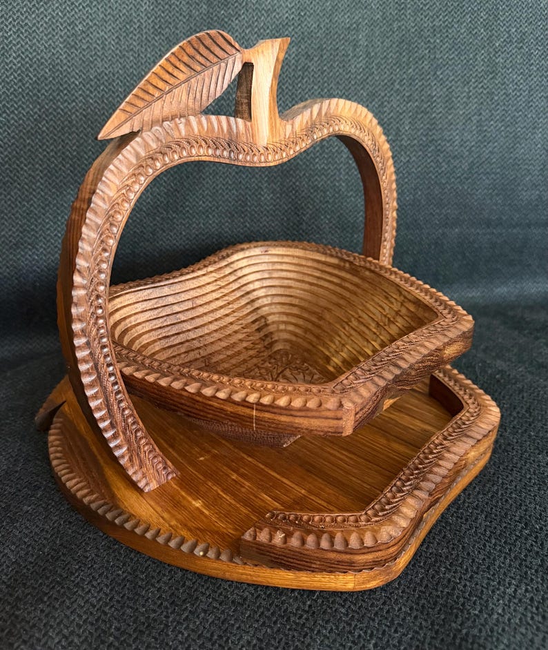 May include: A wooden apple-shaped basket with a carved leaf detail. The basket has a layered design and sits on a matching base. The wood has a warm, natural tone, and the basket is likely used for decorative or functional purposes.