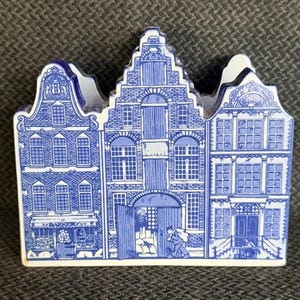 May include: A blue and white ceramic decorative piece shaped like three Dutch houses. The houses feature detailed architectural designs, including windows, doors, and brickwork. The piece is likely a souvenir or decorative item.