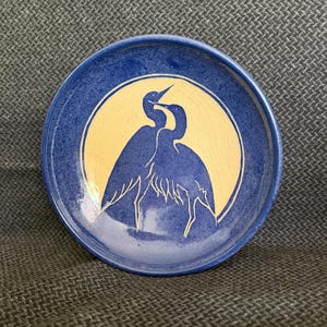May include: A round, blue ceramic plate with a cream-colored center featuring a silhouette of two birds. The birds are facing each other, with their beaks touching. The plate has a textured surface and a raised edge.