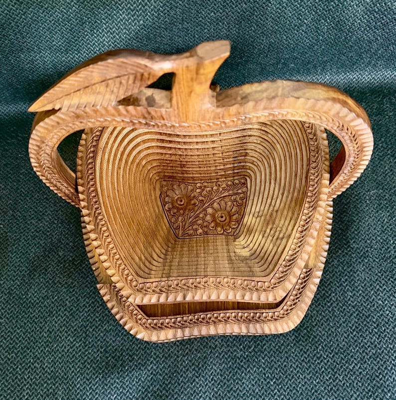 May include: A handcrafted wooden basket with a unique apple shape. The basket features intricate carved details, including a floral design on the base and a leaf-shaped handle. The natural wood tones create a warm, rustic aesthetic.