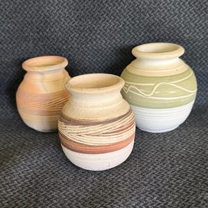 May include: Three small ceramic vases with rounded bodies and flared rims. The left vase is light tan with horizontal etched lines. The center vase features bands of brown, tan, and white with textured grooves. The right vase is sage green and white.