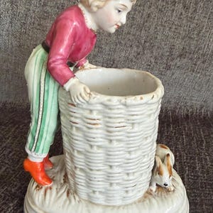 May include: A porcelain figurine of a blonde child wearing a red shirt and green striped pants, leaning over a white cylindrical basket-weave container. A small brown and white rabbit sits on the base next to the basket on a textured surface.