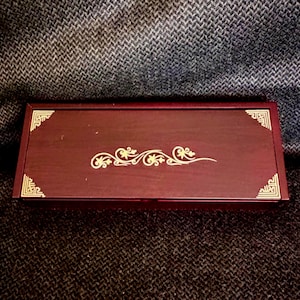 May include: A rectangular, dark red wooden box with gold-colored decorative accents. The box features ornate corner designs and a central floral scroll pattern. The box is closed and sits on a textured, dark gray surface.