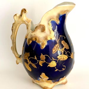 May include: A decorative ceramic pitcher with a dark blue body and a cream-colored rim and base. The pitcher features gold leaf designs of vines and leaves. The handle and accents are also gold. The pitcher has a unique, ornate shape.