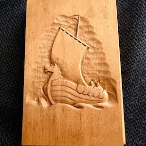 May include: A rectangular wooden carving depicting a Viking ship with a dragon head at the bow. The ship has a large sail and is carved in a light brown wood. The carving is detailed, showing the waves and the ship's design.