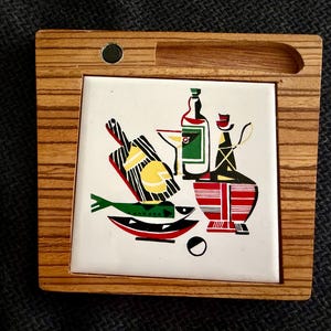 May include: Square wooden trivet with a colorful retro design. The ceramic tile features a still life of bottles, a fish, and a bowl. The trivet has a built-in handle and a small magnet.