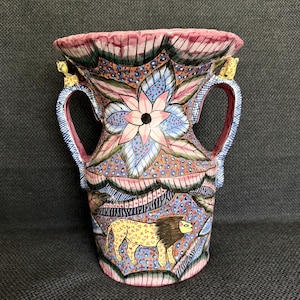 May include: A ceramic vase with two handles featuring a hand-painted design of a lion, floral motifs, and abstract patterns in shades of pink, blue, yellow, and black. The rim is scalloped, and small animal figures are attached to the handles.