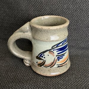 May include: A speckled ceramic mug with a light beige crackle glaze finish. The mug features a hand-painted graphic of a fish with blue, orange, and black details. It has a thick, curved handle and a flared base, showcasing a rustic aesthetic.