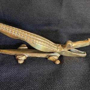 May include: A brass nutcracker shaped like an alligator with a textured, scaled back and an open mouth. The metal tool features a hinged design that allows the upper jaw to move, revealing a serrated interior surface for gripping nuts.