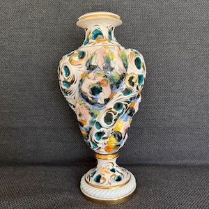 May include: A white ceramic vase featuring intricate openwork cutouts and ornate scrollwork. The surface is decorated with hand-painted floral motifs in shades of pink, green, yellow, and black, accented with gold trim along the rim and base.