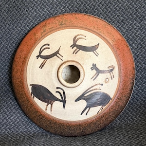 May include: A round ceramic vessel with a central opening, featuring a wide, textured terracotta-colored rim and a cream-colored center decorated with four hand-painted black animal figures, including three horned goats and one small canine.