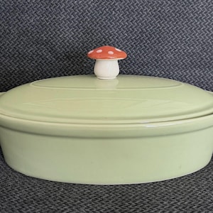 May include: A light green ceramic casserole dish with a lid and two handles. The lid is topped with a mushroom-shaped knob with a red and white spotted cap. The dish is oval-shaped.