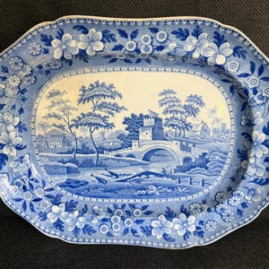 May include: An oval ceramic platter featuring a blue and white transferware pattern. The center depicts a landscape with a stone bridge, a castle, and trees. The wide rim is decorated with a dense floral motif of blossoms and leaves.