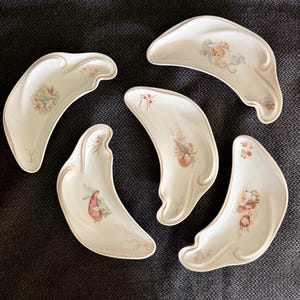 May include: A set of five crescent-shaped white ceramic dishes, each featuring unique, delicate illustrations of seashells, coral, and marine life in soft pastel tones, with subtle gold-toned trim along the curved, scalloped edges.
