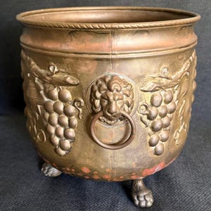 May include: A round copper planter featuring embossed grape clusters and foliage on the exterior. The vessel includes two lion head handles with attached rings and stands on three claw-shaped feet. The metal surface shows a warm patina.