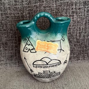 May include: A ceramic wedding vase with a teal and cream gradient finish. The vessel features a central handle and black line art depicting a teepee, a mountain range, and a stepped structure. A sticker reads: MADE IN ARIZONA BY ROSEMARY'S CERAMICS.