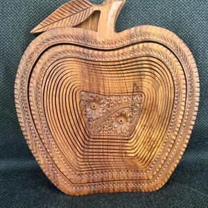 May include: Wooden apple-shaped basket with intricate carved details. The basket features a leaf and stem design, with a floral pattern carved into the center. The basket is made of light brown wood and has a collapsible design.