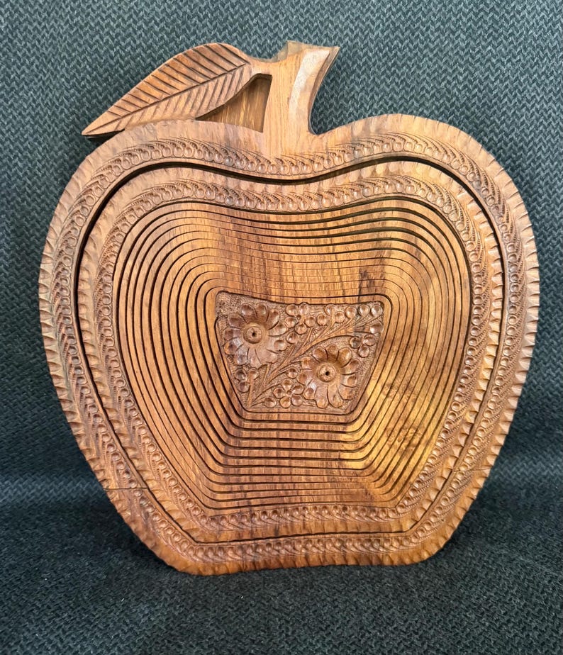 May include: Wooden apple-shaped basket with intricate carved details. The basket features a leaf and stem design, with a floral pattern carved into the center. The basket is made of light brown wood and has a collapsible design.