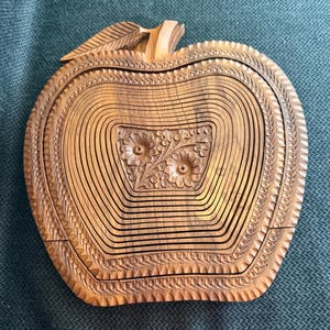 May include: A wooden apple-shaped basket with intricate carved details. The basket is made of light brown wood and features a floral design in the center. The edges are decorated with a repeating pattern. The basket is designed to expand and collapse.