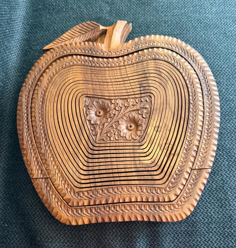May include: A wooden apple-shaped basket with intricate carved details. The basket is made of light brown wood and features a floral design in the center. The edges are decorated with a repeating pattern. The basket is designed to expand and collapse.