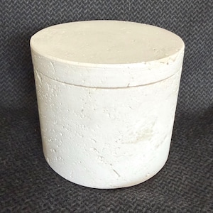 May include: A white, cylindrical container with a matching lid. The container has a textured surface and a rounded base. The lid sits flush with the top of the cylinder. The item is made of a solid, opaque material.