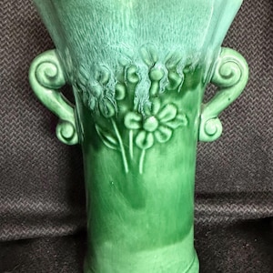 May include: A green ceramic vase with a flared top and two scroll handles. The vase features embossed floral designs and a textured finish. The color gradient transitions from a darker green at the base to a lighter shade at the top.