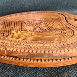 May include: A wooden, collapsible fruit basket with a natural finish. The basket is carved with a floral design in the center and a decorative border. The basket is approximately 10 inches in diameter.